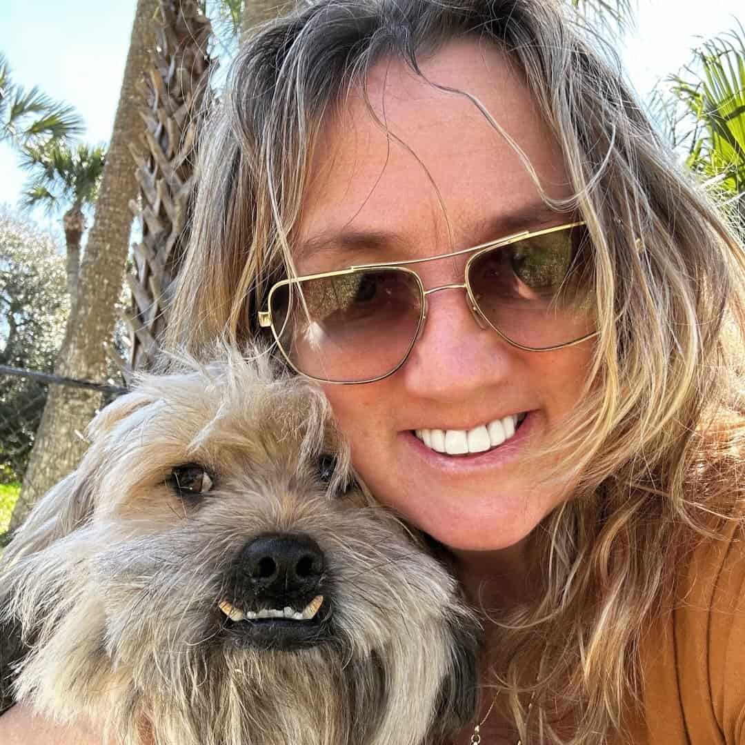 Woman smiling with a fluffy, small dog outdoors in sunny setting; they pose close together for a selfie, palm trees in background.