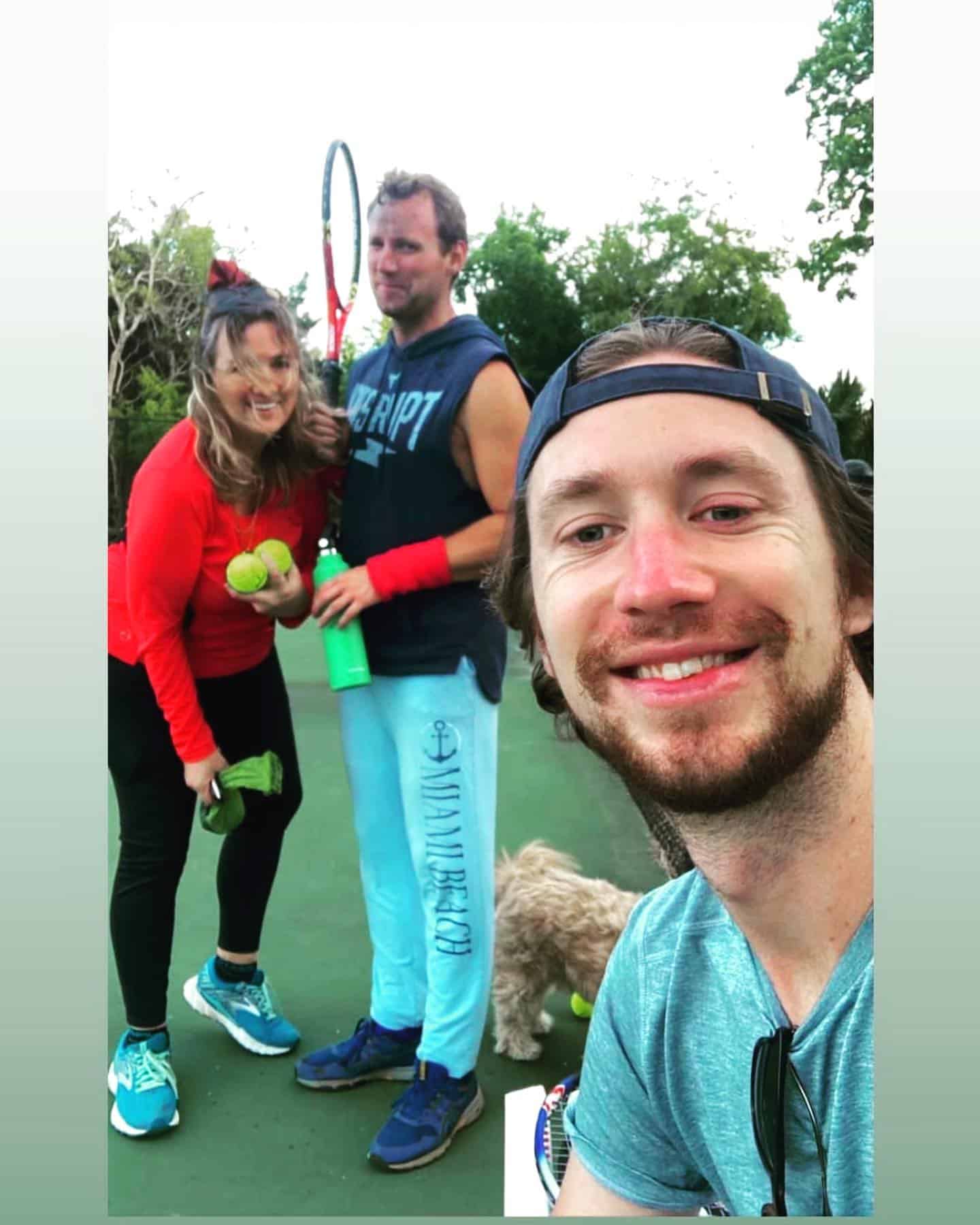 Group of friends on a tennis court posing for a selfie: man with a racquet in the background, woman holding tennis balls, and a man in the foreground taking the photo with a cap on backward; a small dog nearby.
