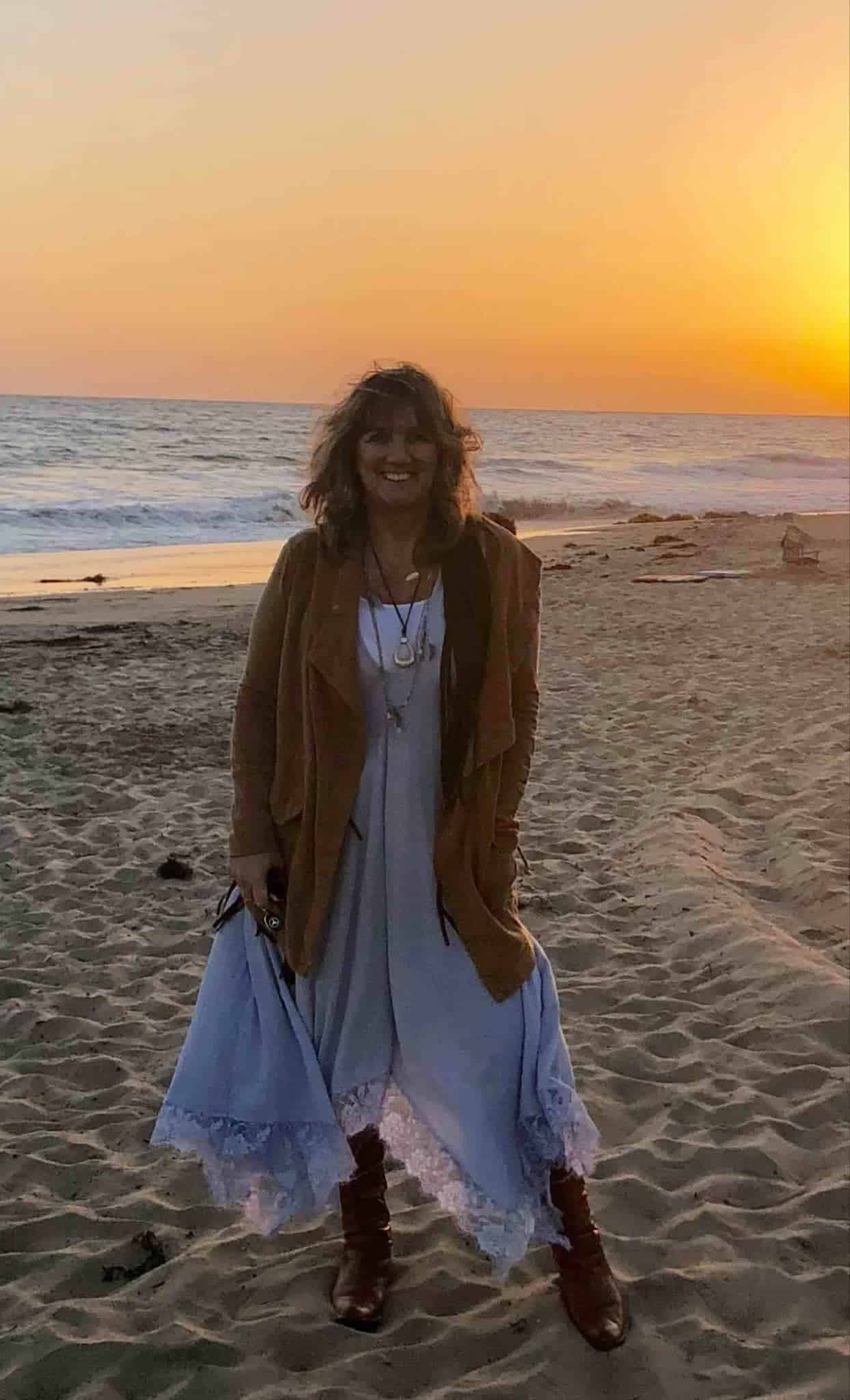 Woman standing on a sandy beach at sunset, wearing a brown jacket over a light dress and brown boots, smiling at the camera.