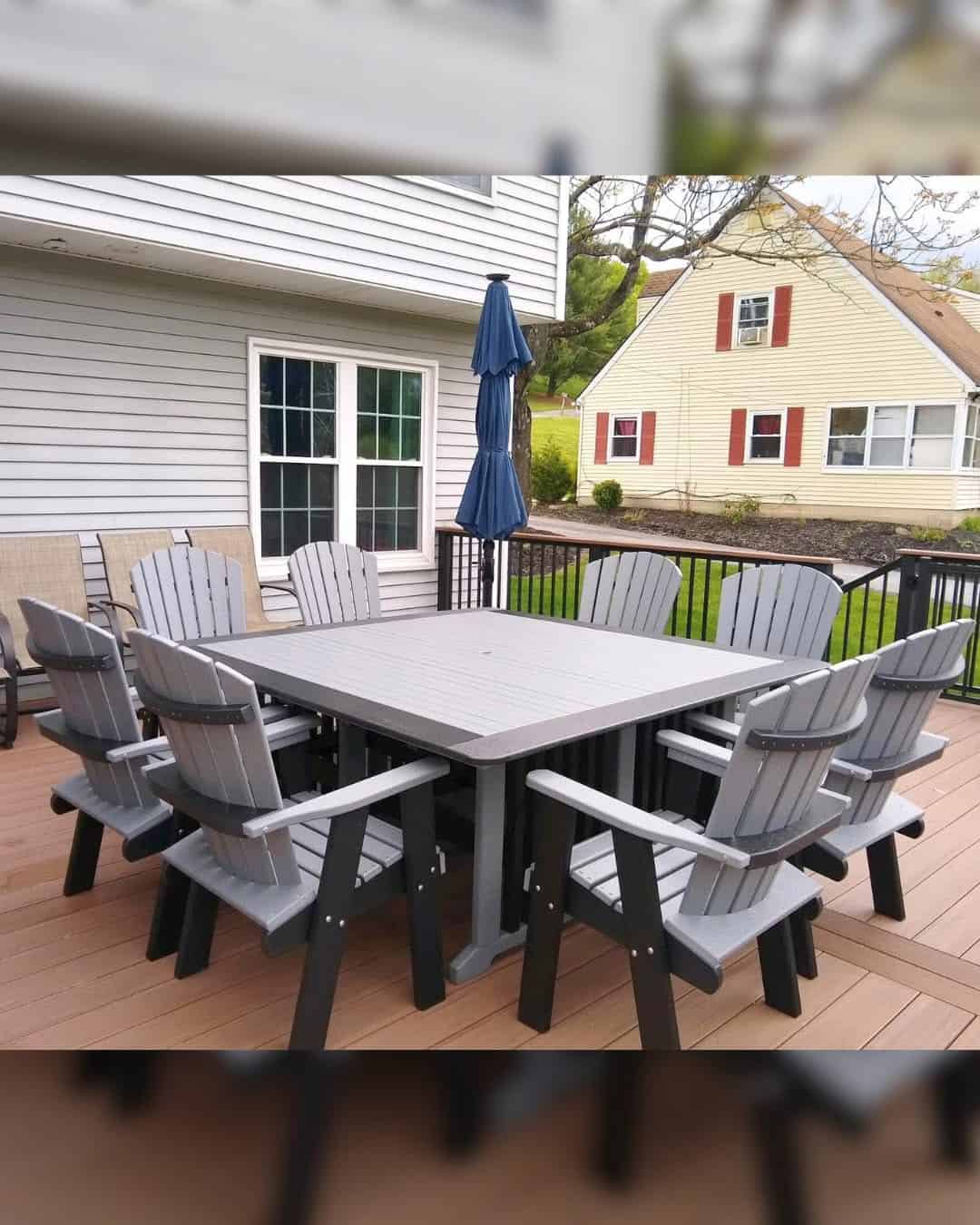 Large gray outdoor dining table with twelve matching Adirondack chairs on a wooden deck, blue umbrella folded nearby.