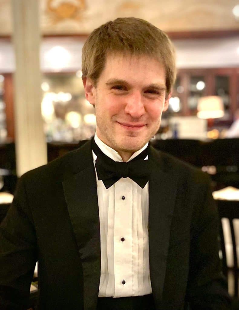 Young man in a black tuxedo and white bow tie, smiling for the camera indoors at a formal event