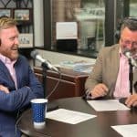 Two men wearing headphones and speaking into microphones at a podcast table, papers and a polka-dot cup in view.