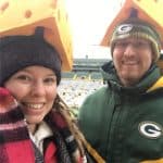 Smiling couple wearing cheesehead hats at a Green Bay Packers stadium, taking a selfie in winter coats and scarves.