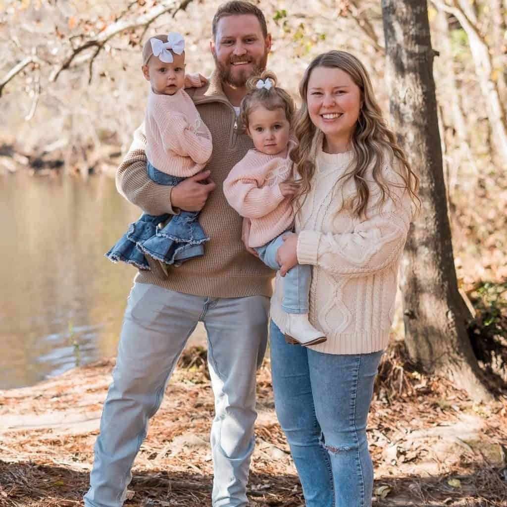 Family of four posing outdoors by a lake in autumn, with mom and dad holding their two young daughters
