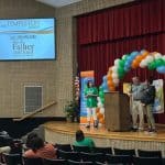 Stage in a school auditorium with a blue screen showing 'Templeton Group' and a balloon arch on the right; people at a podium speak to an audience.