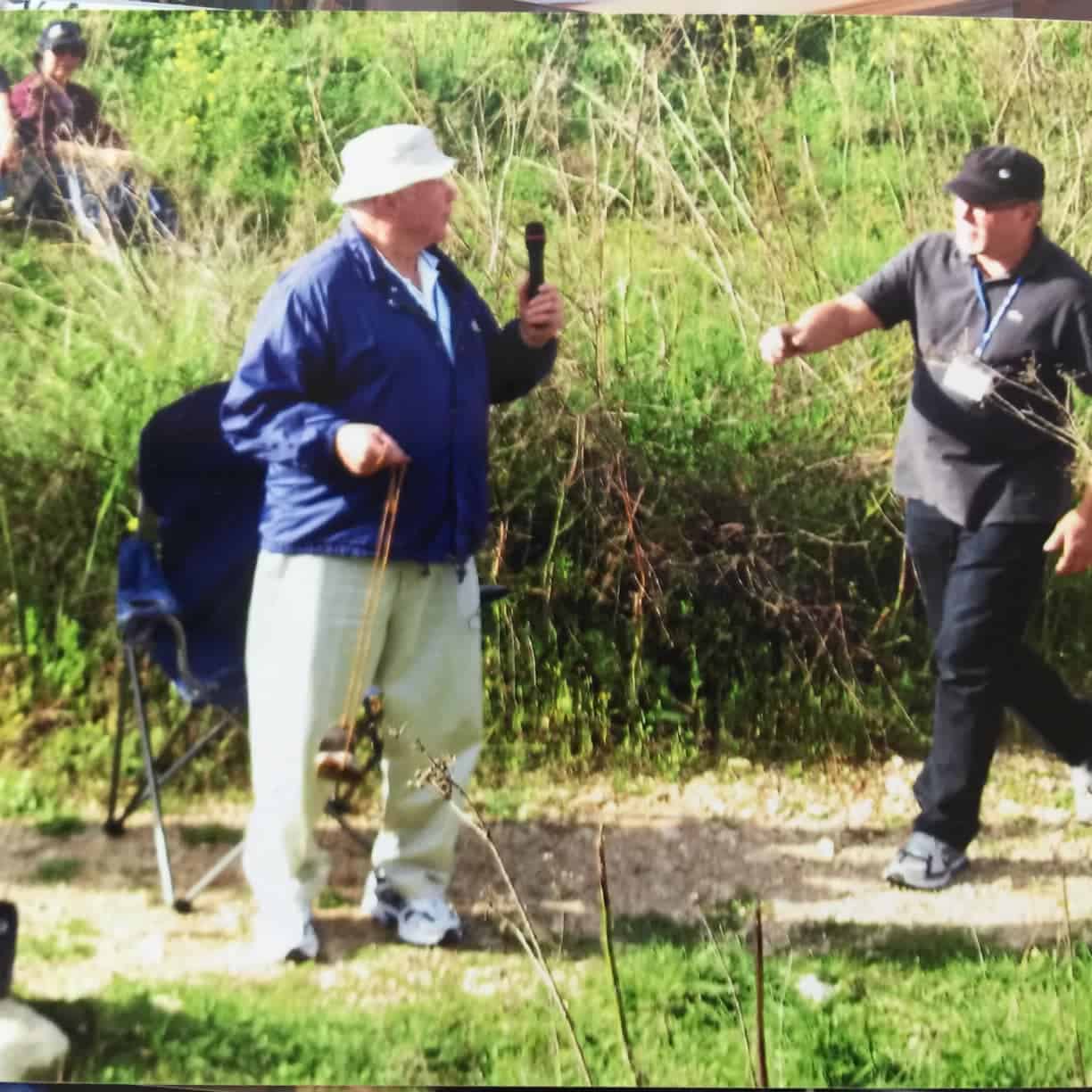 Man in a white hat and blue jacket speaks into a handheld microphone outdoors while another man in a black shirt and cap gestures nearby.