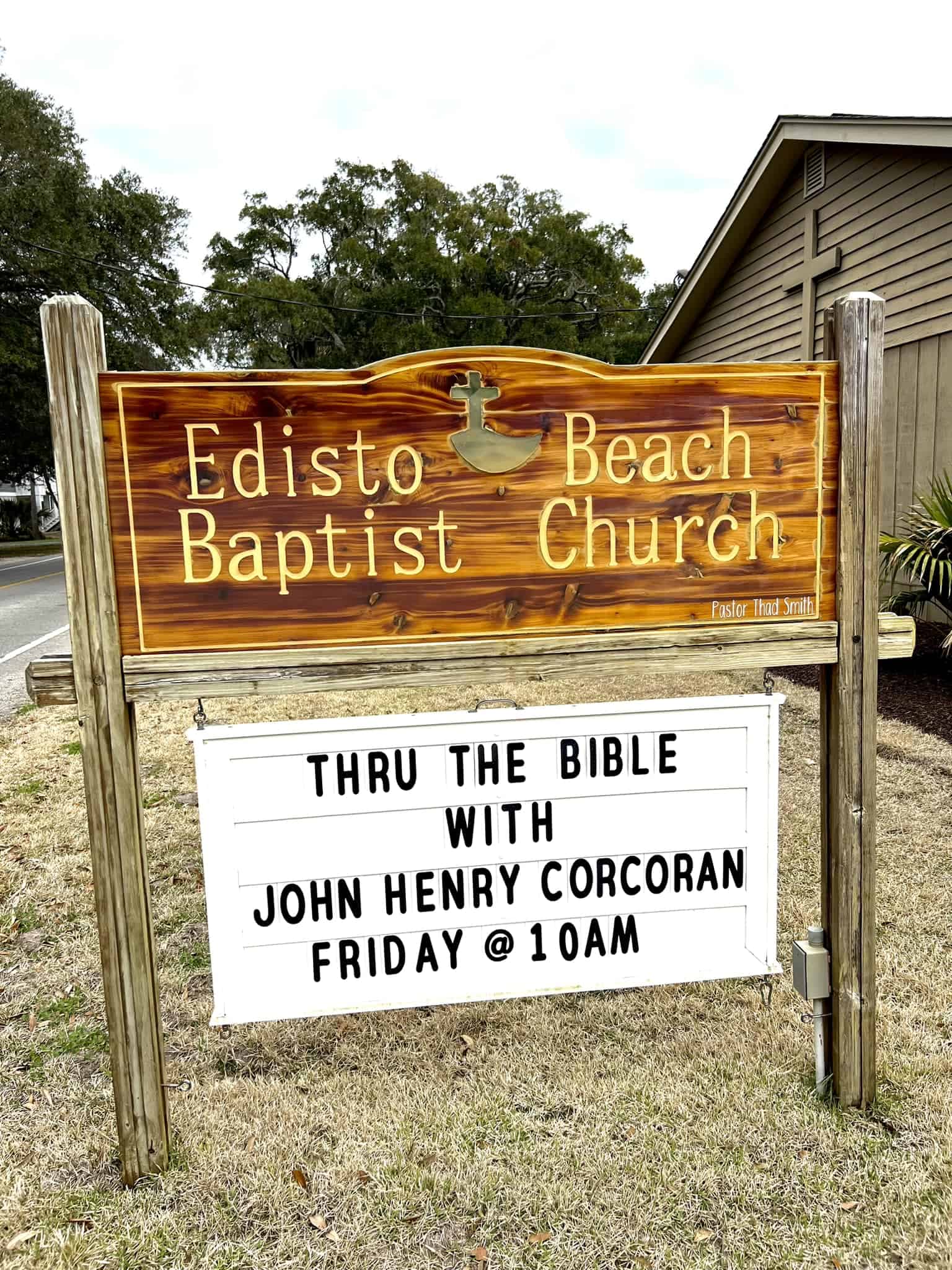 Wooden church sign for Edisto Beach Baptist Church with cross emblem and service text displayed on a sunny day next to a building