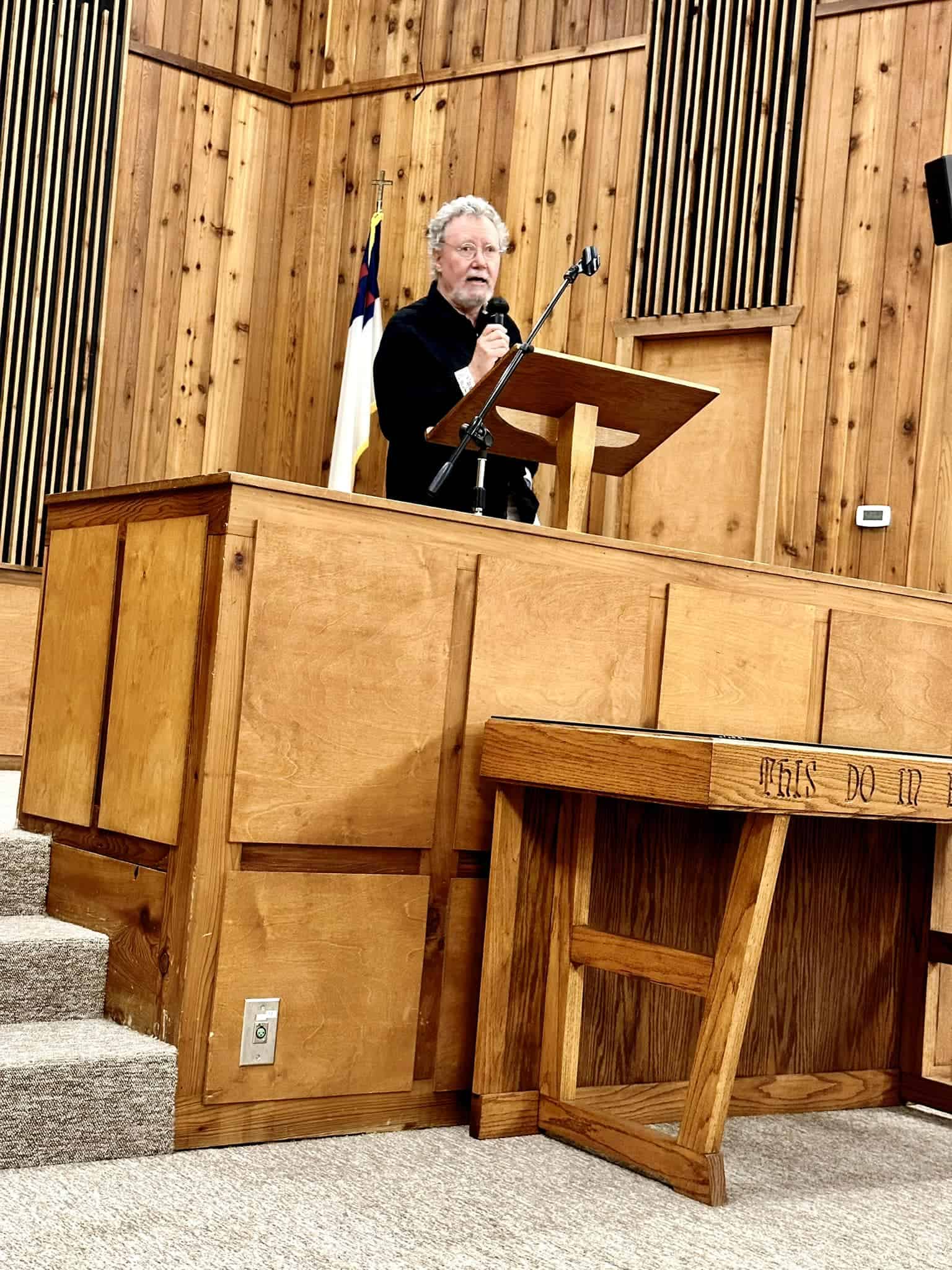 Older man delivering a sermon from a wooden lectern in a church, microphone in hand, flag behind him.