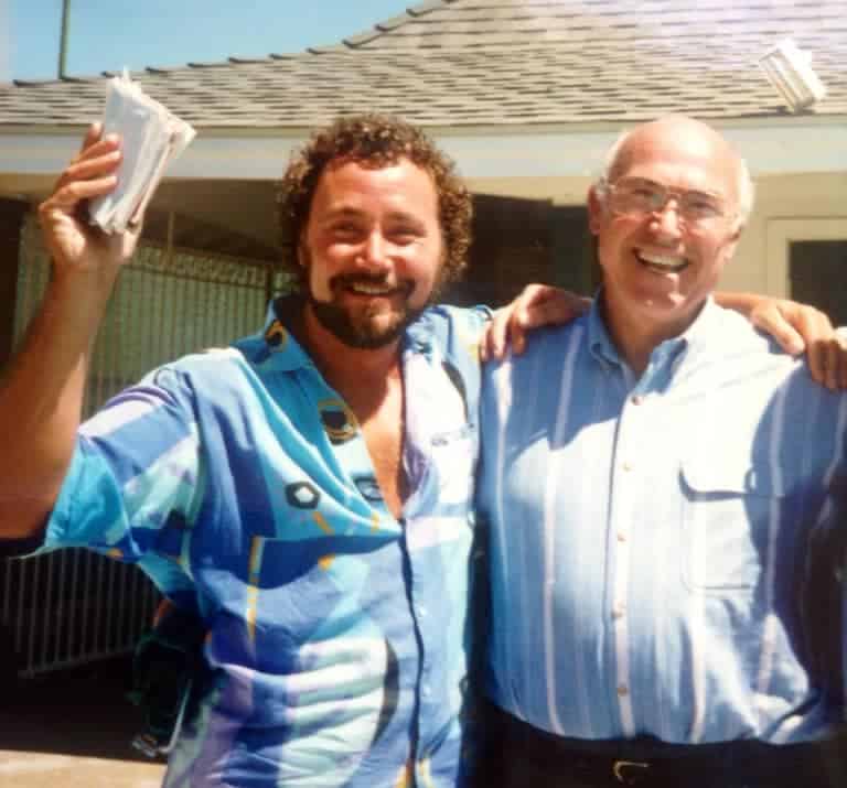Two men outdoors in front of a house, smiling with arms around each other; the man on the left holds a stack of papers.