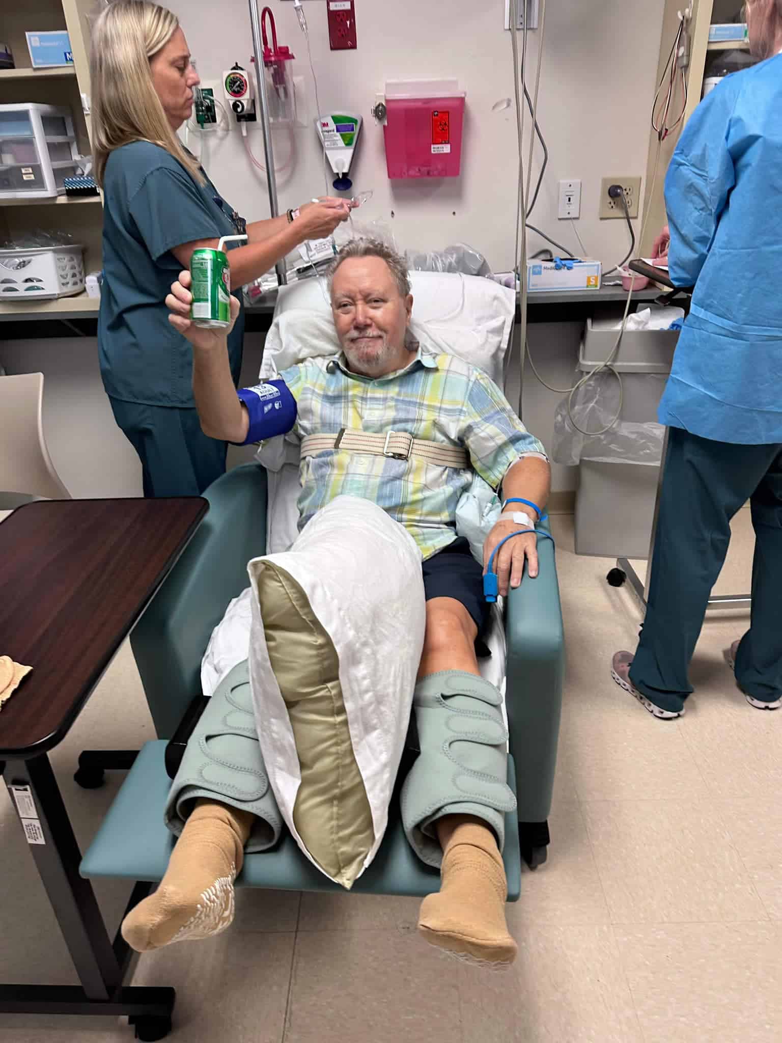 Older man in a plaid shirt reclining in a hospital chair, donating blood with an IV in his arm; nurse in teal scrubs attends to him while another clinician stands nearby, and he holds a green can in his left hand; his leg is in a brace.