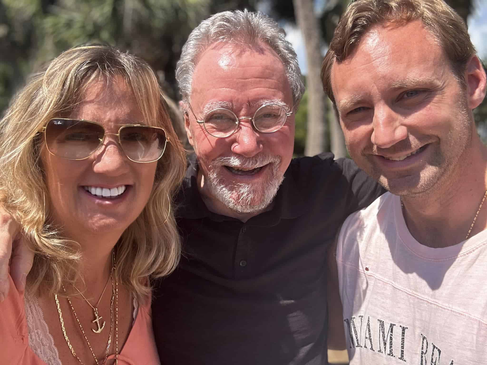 Three adults smiling in a sunny outdoor selfie: a woman with sunglasses on the left, a gray‑haired man in the middle, and a man in a light shirt on the right.