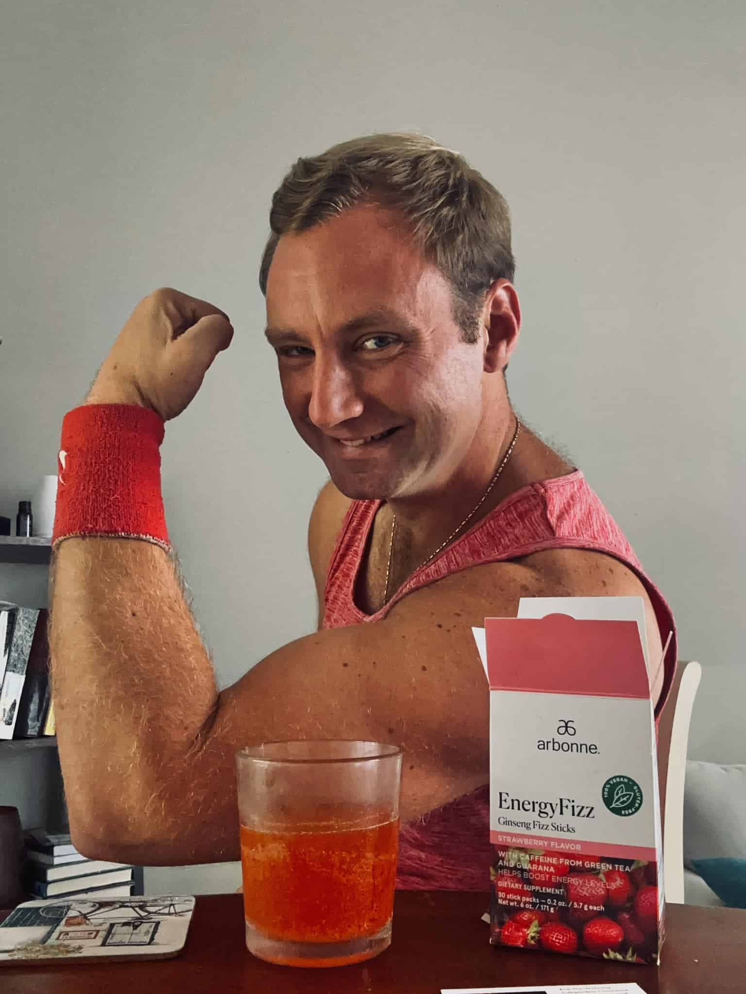 Smiling man in a red tank top flexing his right bicep, with an orange drink and Arbonne EnergyFizz box on a table in front of him.