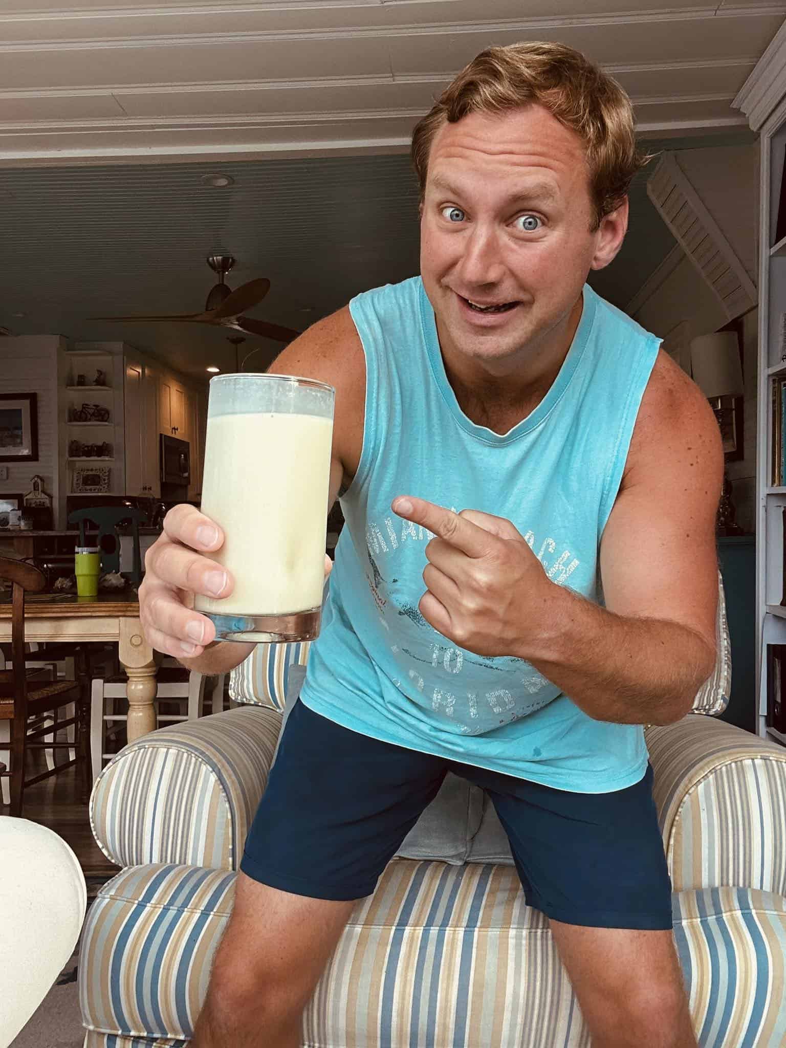 Man in a turquoise tank top posing with a tall glass of milkshake, pointing at it in a bright indoor living space.