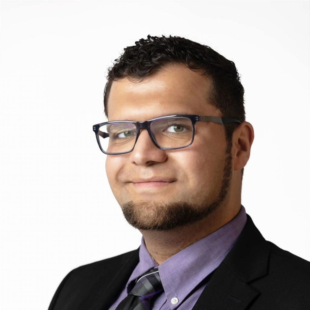 Portrait of a man in a dark blazer, purple shirt, and striped tie, smiling slightly with glasses.