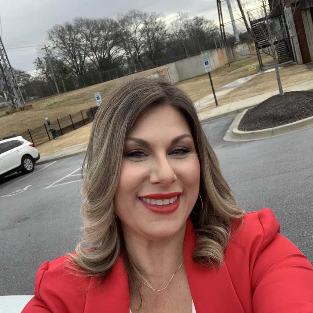 Close-up of a smiling woman wearing a red blazer taking a selfie in a parking lot with bare trees in the background.
