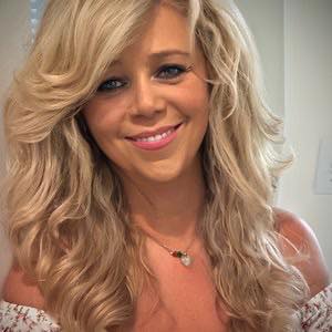 Portrait of a smiling woman with long wavy blonde hair and a necklace, looking at the camera.
