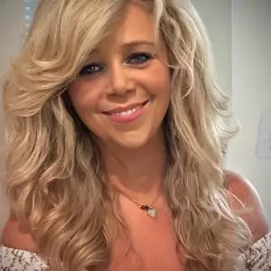 Portrait of a smiling woman with long wavy blonde hair and a necklace, looking at the camera.
