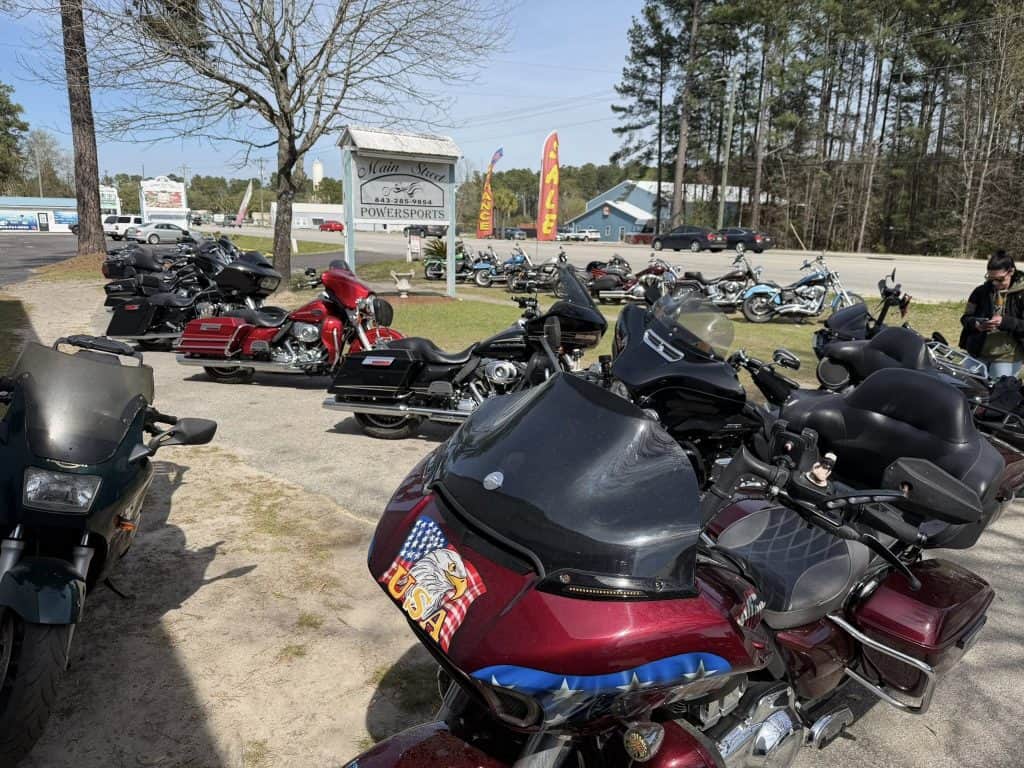 Outdoor motorcycle dealership lot with numerous touring bikes and a 'Main Street Powersports' sign visible in the background, plus sale banners nearby.