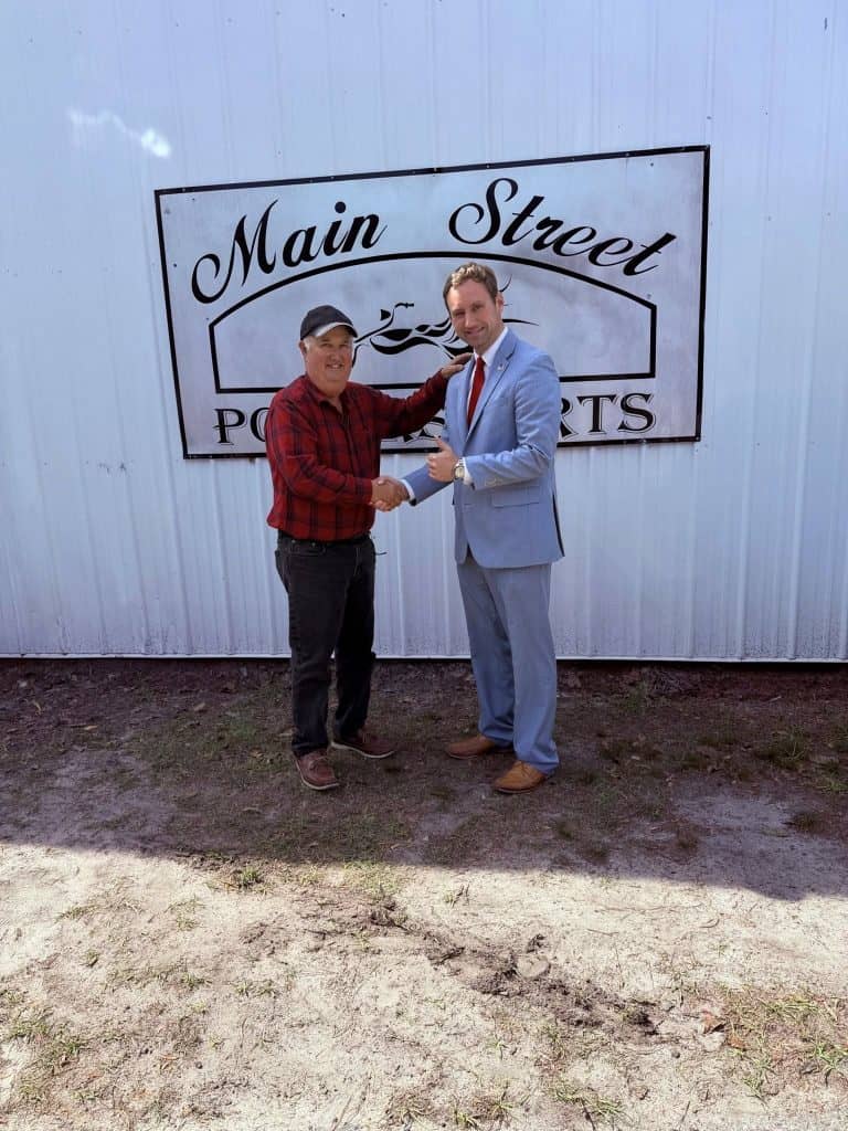 Two men shake hands in front of a Main Street sign on a light blue metal wall; one wears a red plaid shirt and cap, the other a light blue suit with a red tie.