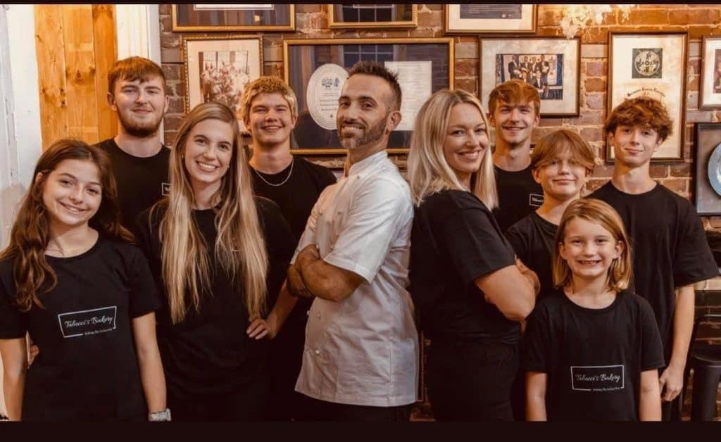 Group of bakery staff posing for a team photo in a brick-walled shop; two adults center (man in white chef coat and woman in black) flanked by several smiling teammates in black uniforms, framed awards on the wall behind.