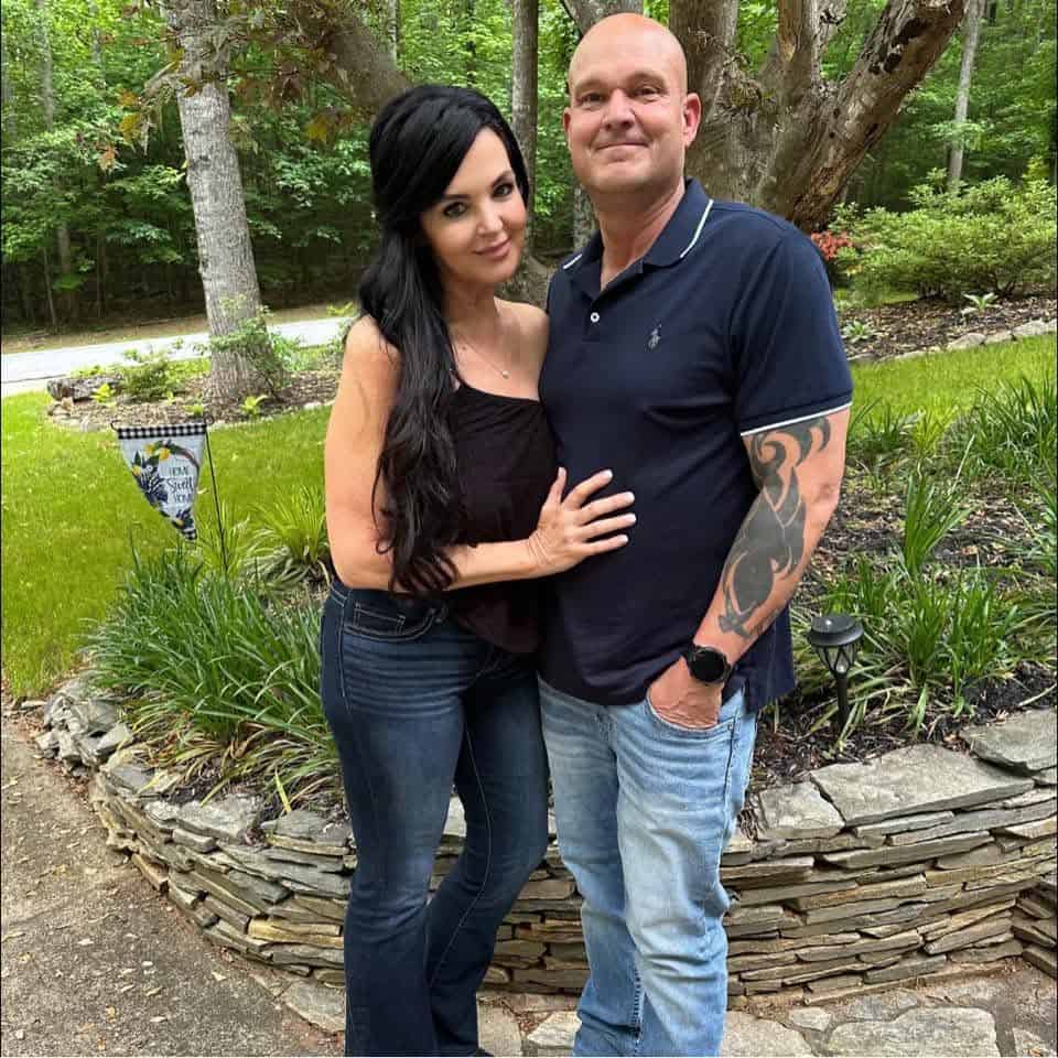 Couple standing close together outdoors in a garden, woman with long black hair wearing a black top and jeans, man in a navy polo and jeans with a tattooed arm.