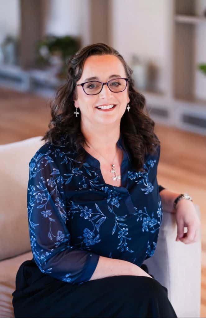 Smiling woman with glasses in a blue floral blouse seated on a sofa in a bright living room.