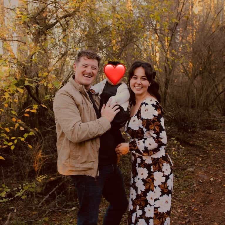 Smiling man and woman hold their baby outdoors in autumn; the baby's face is covered with a heart emoji.