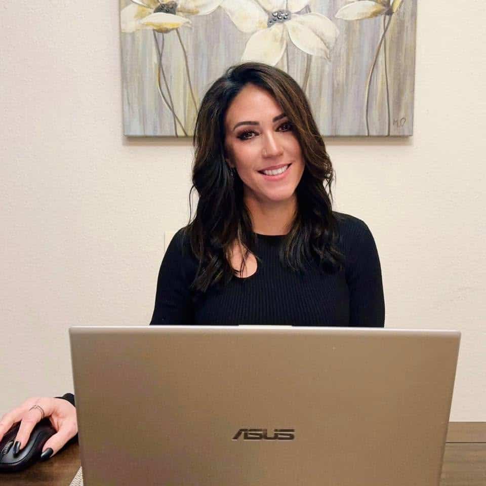 Smiling woman with dark hair in a black top sits at a desk with an ASUS laptop and a wireless mouse in front of her.