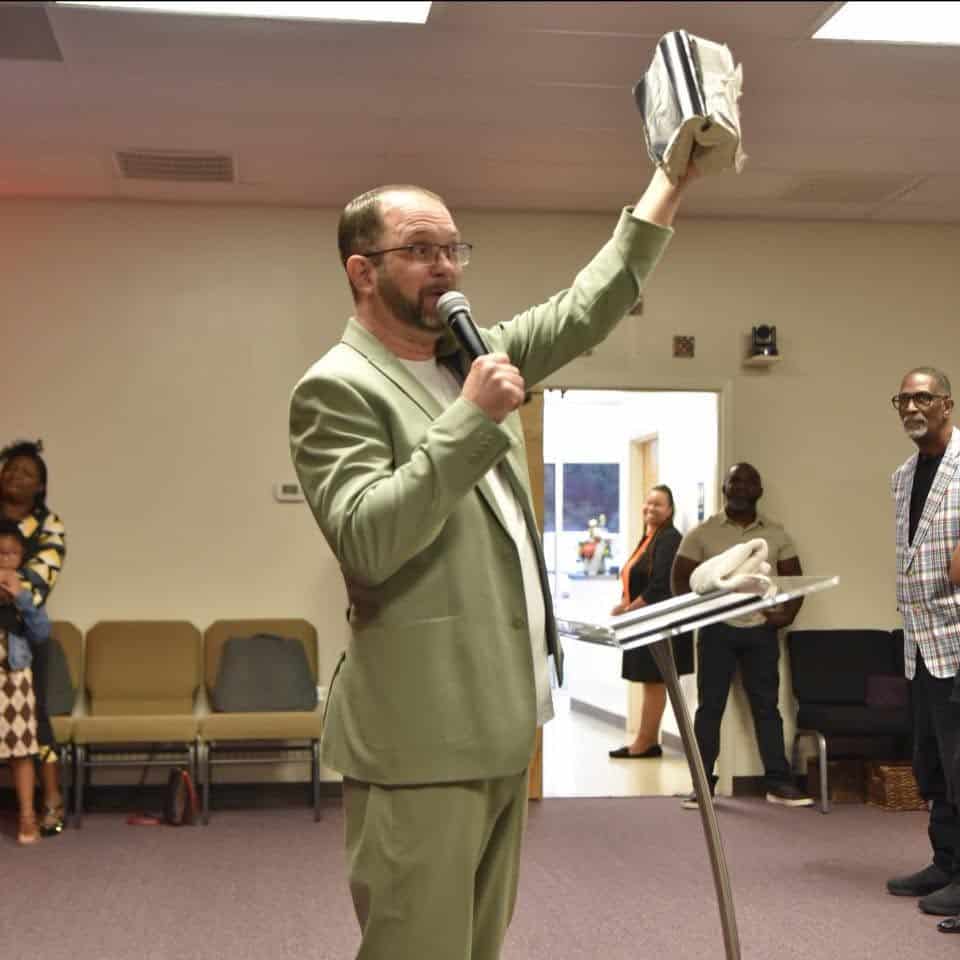 Man in a light green suit raises a wrapped object while speaking into a microphone at a podium in a meeting room.