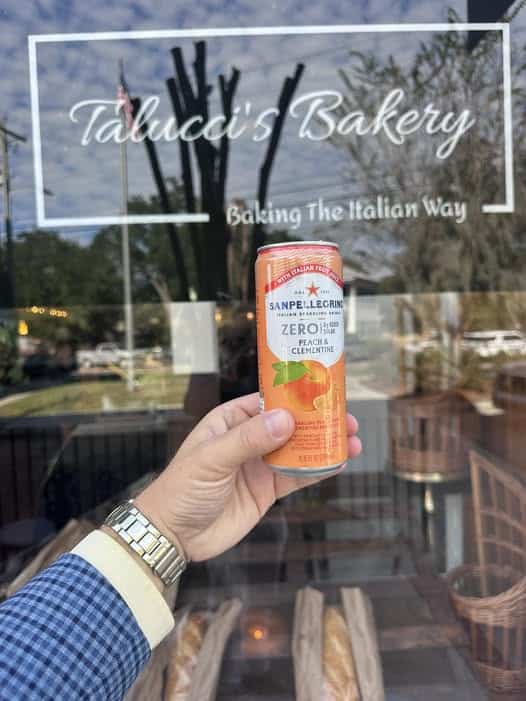 Hand holding an orange San Pellegrino can in front of a bakery window with 'Tabuchi's Bakery' sign