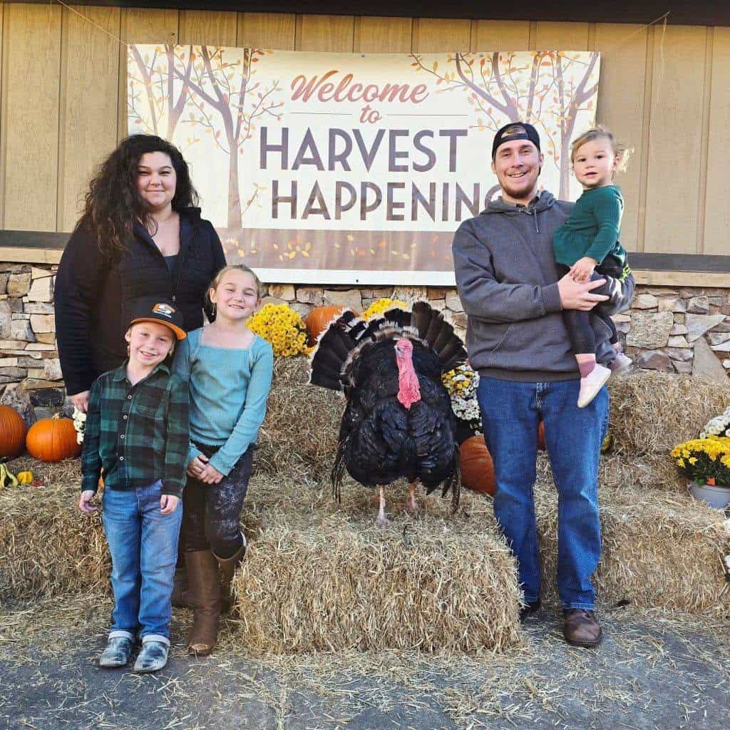 Family of five posing with a large turkey on hay bales at a fall harvest festival; sign reads 'Welcome to Harvest Happening'.