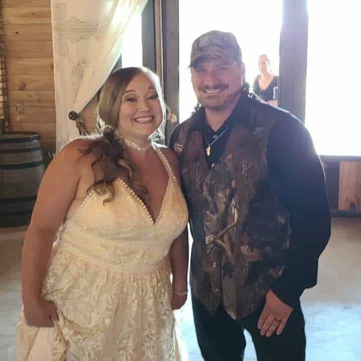 Smiling couple posing for a photo at a wedding venue; woman in ivory lace dress with pearls, man in camouflage vest and cap.