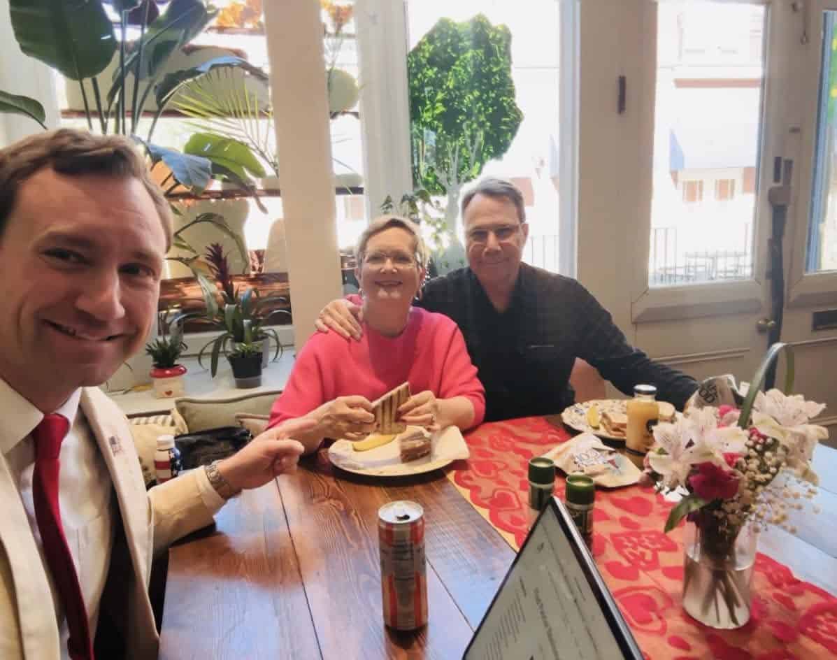 Three people sit at a wooden dining table in a bright room, smiling at the camera during a meal; woman in pink holds a sandwich, man in black sits beside her, friend to the left in a white suit and red tie.