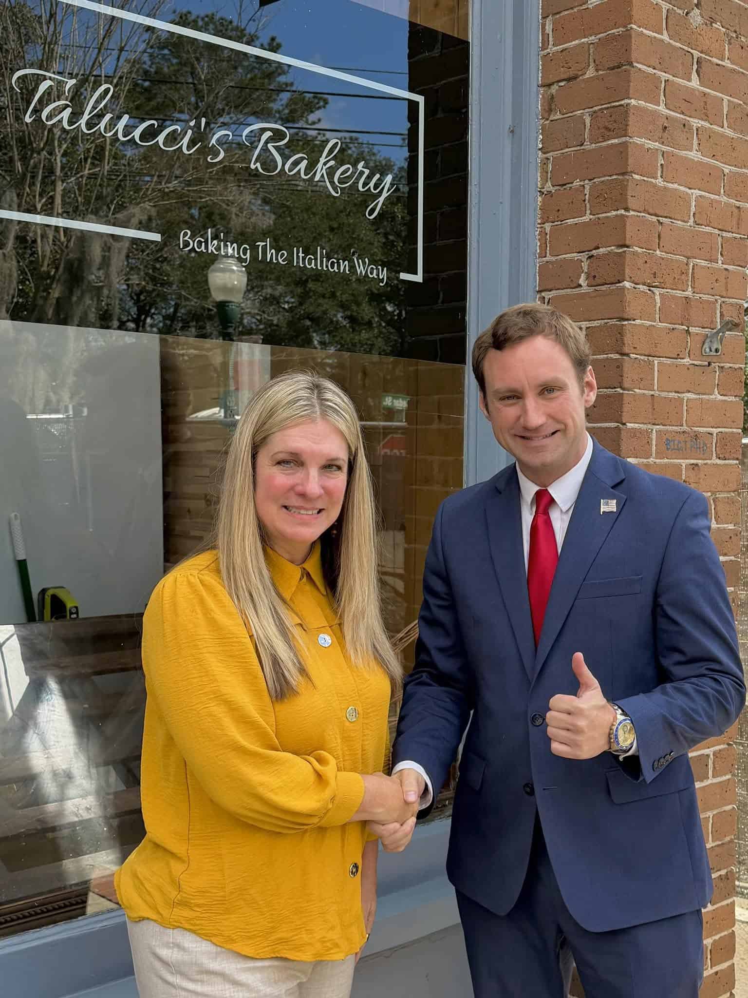 Two people shake hands outside a brick bakery storefront with 'Valucci's Bakery' in the window; woman in yellow top and man in a navy suit give a thumbs-up.