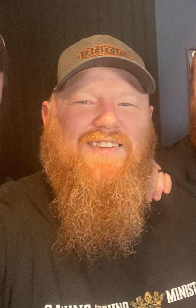 Smiling man with a long red beard wearing a brown cap, dark shirt, and a hand on his shoulder visible at the edge of the frame
