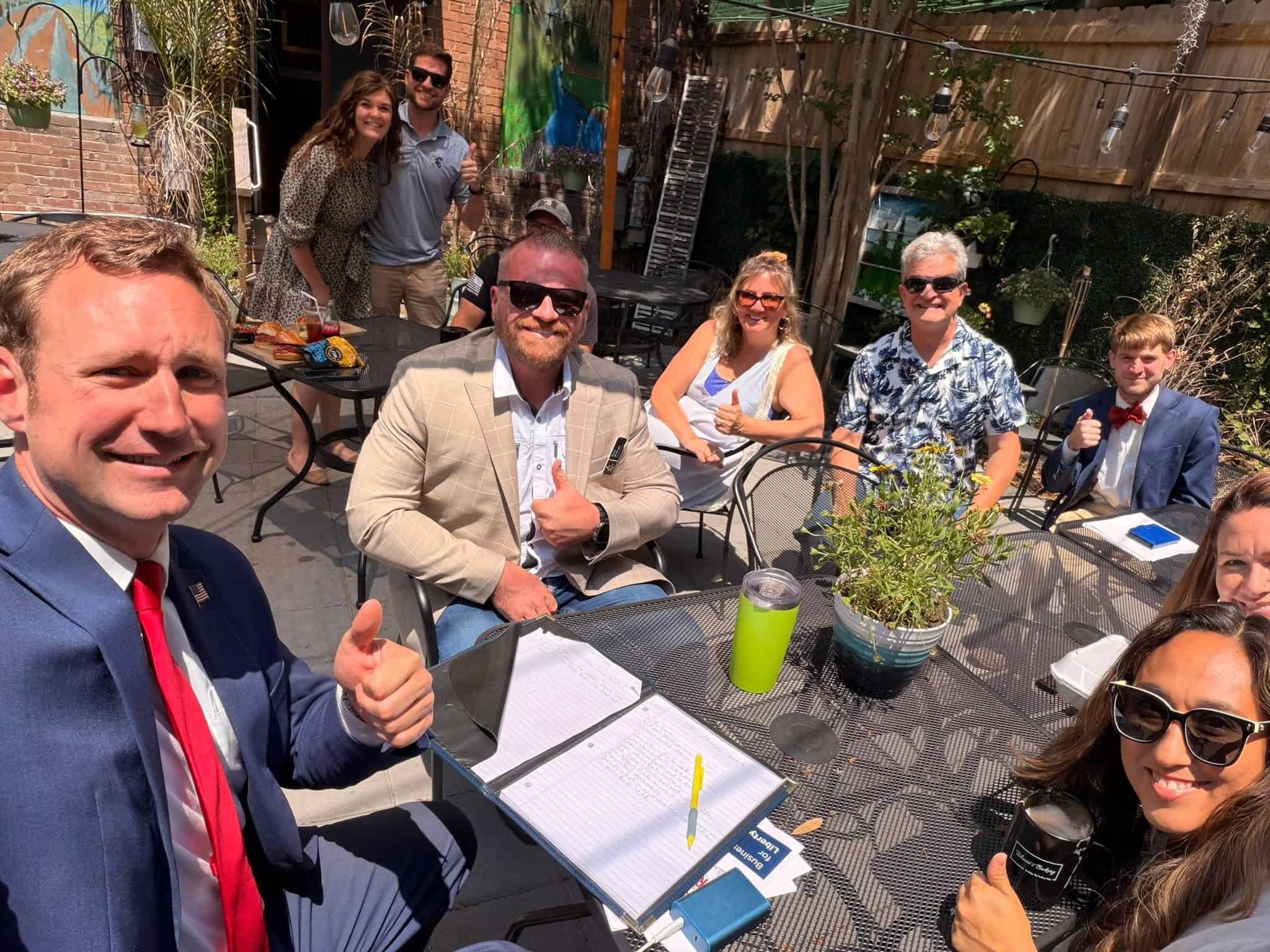 Group of adults in business casual attire around an outdoor cafe table, smiling with documents and drinks, giving thumbs up.