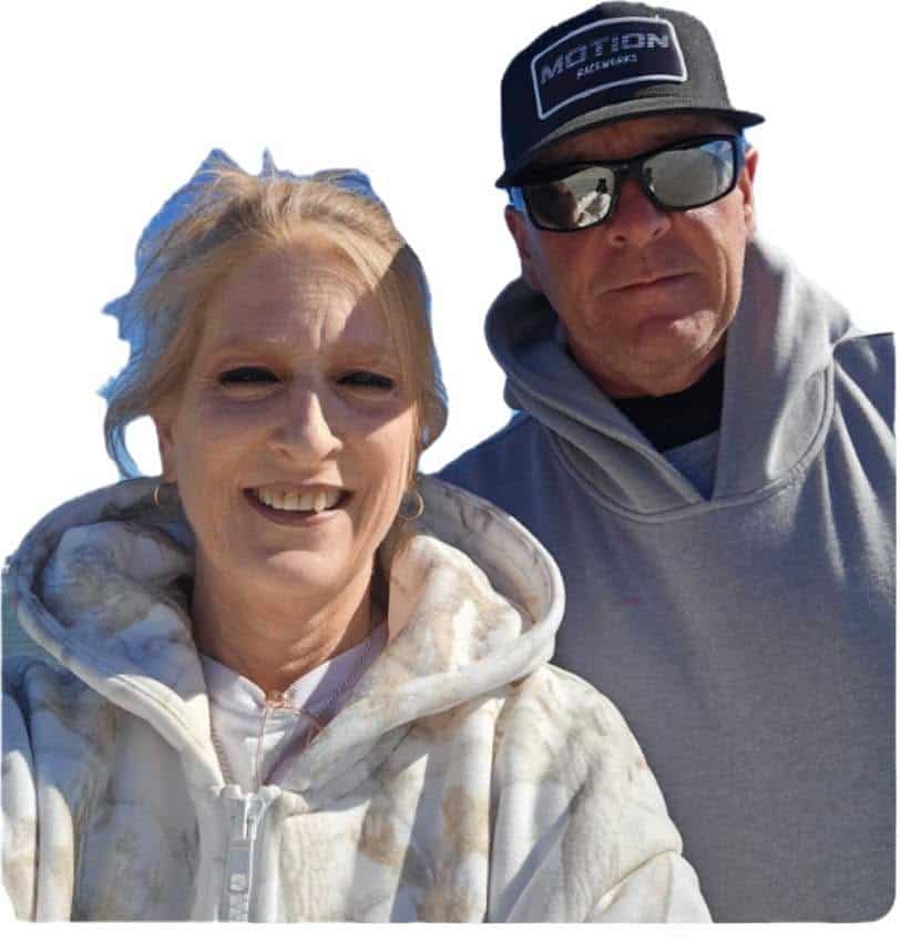 Two adults posing for a sunny selfie outdoors, smiling at the camera, both wearing hoodies.