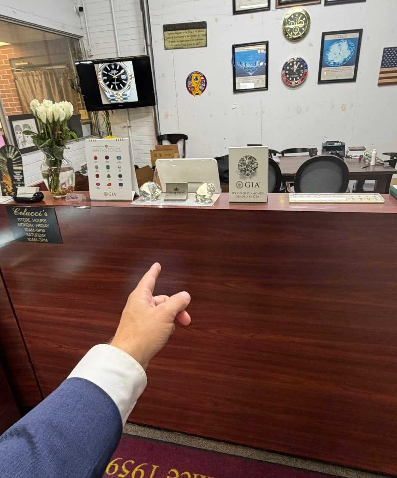 Hand in a blue-suited sleeve points at a jewelry-store reception desk, with clocks and framed wall decor behind it.