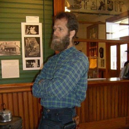 Bearded man in a blue plaid shirt standing with arms crossed beside historical photo displays on a green wall.