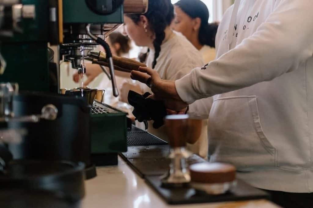 Barista hands pressing a portafilter under an espresso machine in a cafe, with customers in the background.
