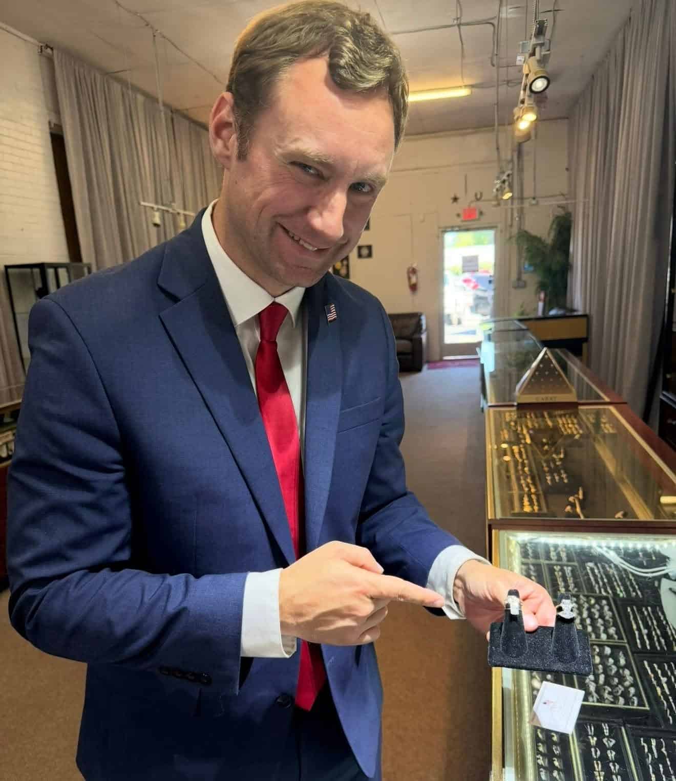 Man in a blue suit and red tie stands at a jewelry display case, pointing to rings inside the glass case in a shop showroom.