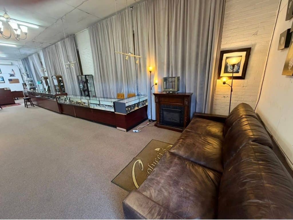 Store interior with long row of glass display cases along the left, and a brown leather sofa on the right; a wooden fireplace and lamps sit near the back.