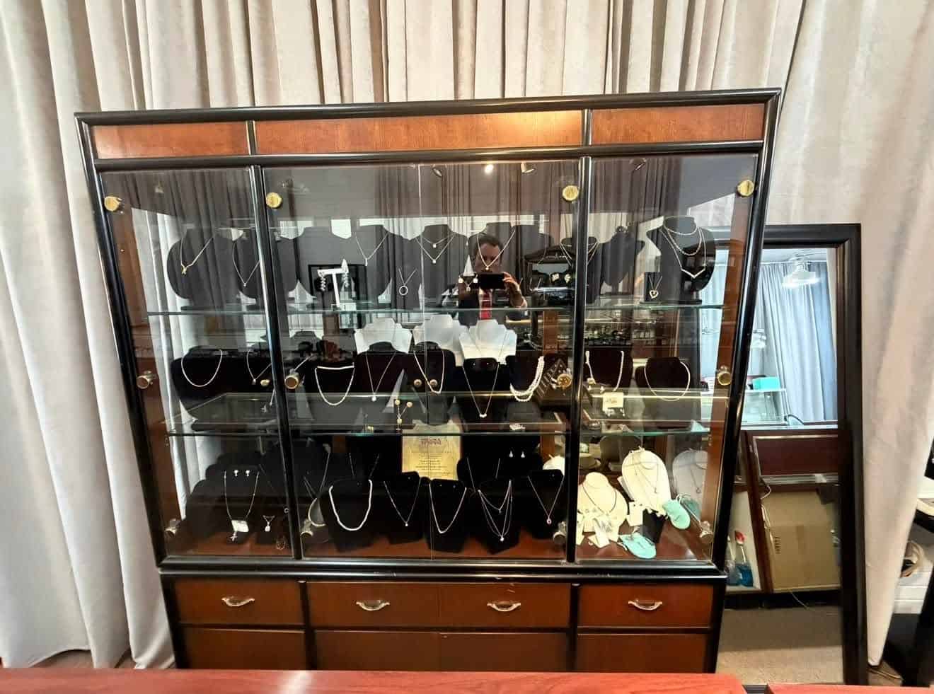 Glass-front jewelry cabinet displaying necklaces on black busts; photographer visible in the glass reflection.