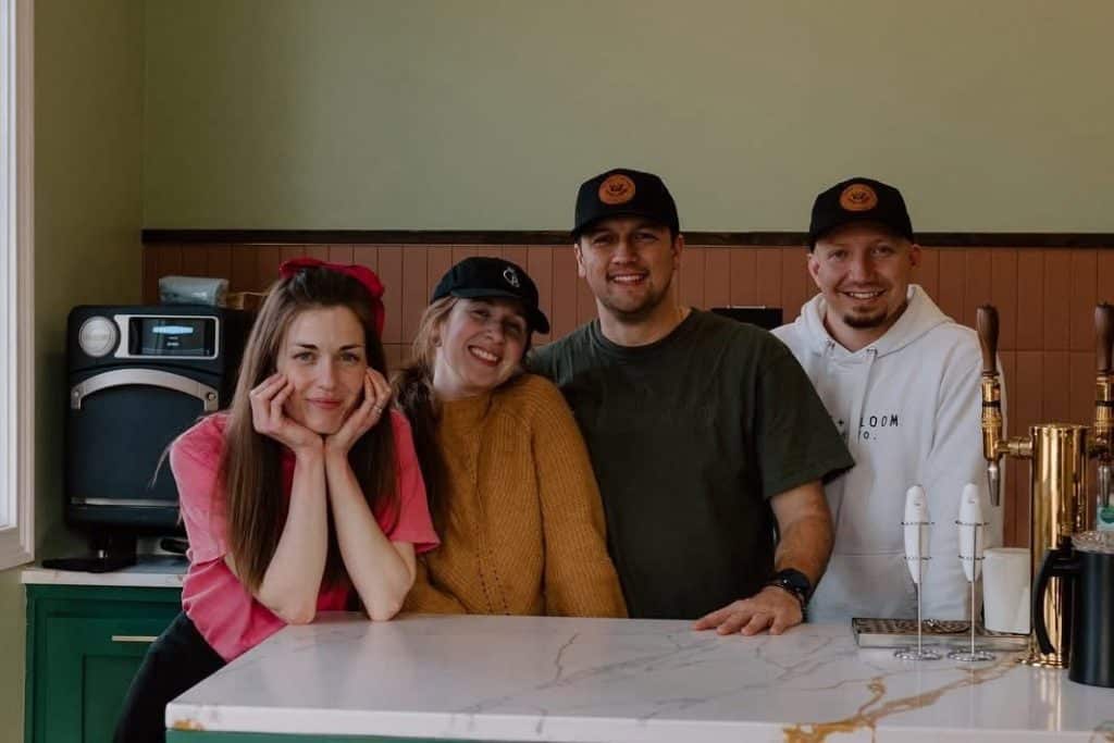 Four friends posing together at a cafe counter, smiling at the camera with a coffee machine and taps visible in the background.