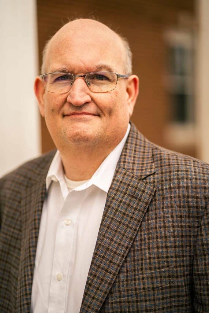 Portrait of an older man with glasses wearing a brown plaid blazer, looking at the camera and smiling softly.