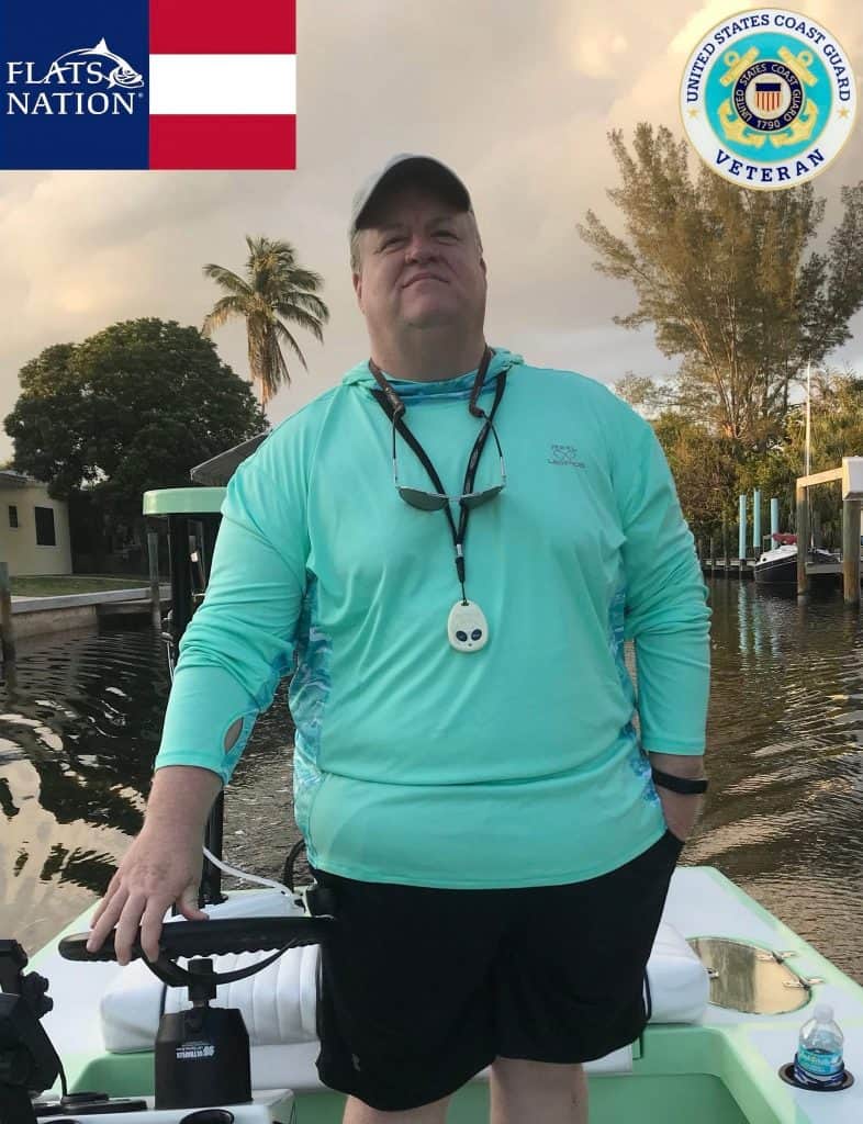 Man wearing a teal long-sleeve shirt standing on a small boat by the water, with palm trees in the background and a lanyard around his neck.