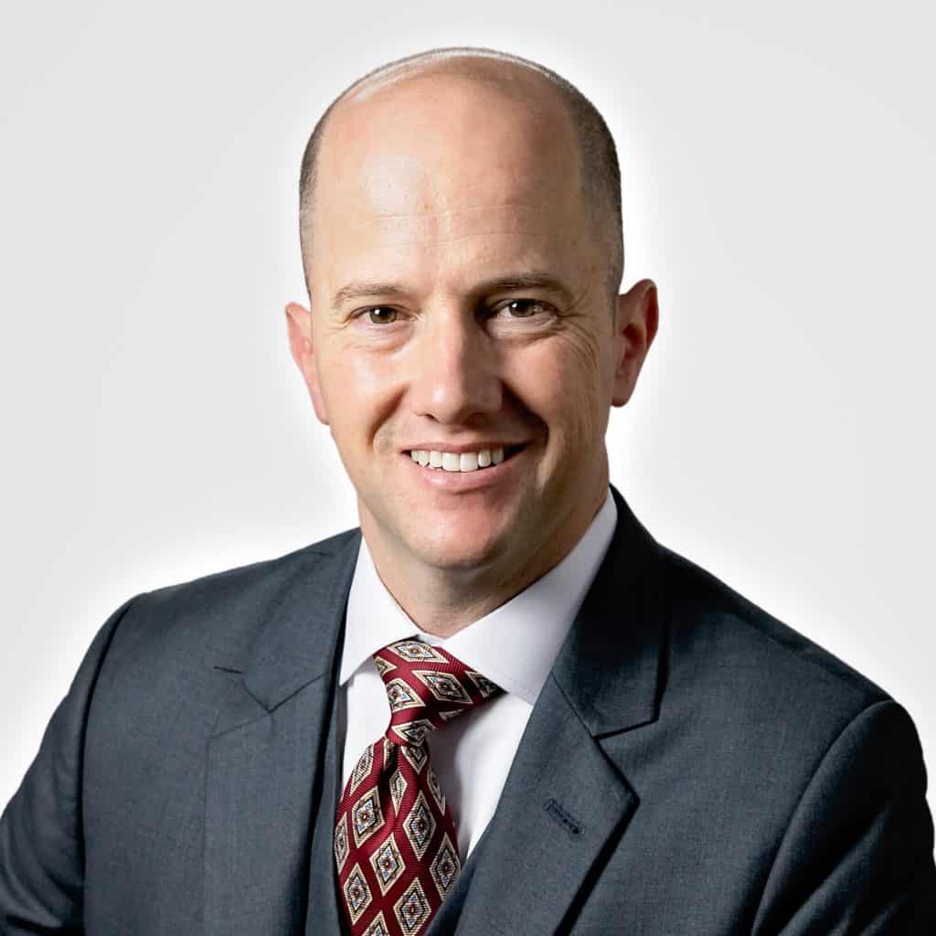 Close-up portrait of a smiling man in a dark suit and patterned red tie against a light background.