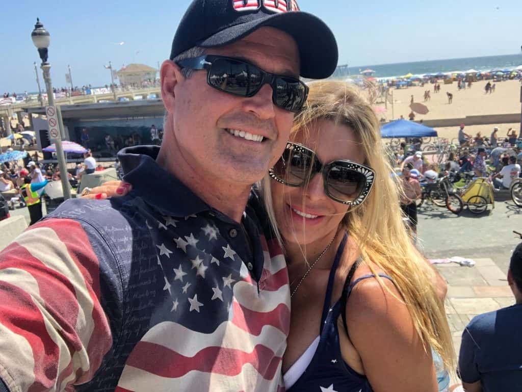 Close-up selfie of a smiling man wearing sunglasses and a cap with a woman at a busy beach boardwalk in the background.
