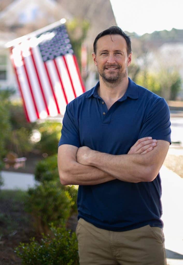 Man in a blue polo shirt standing outdoors with arms crossed, a large American flag in the background.