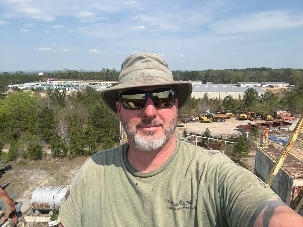 Self-portrait of a man wearing a wide-brimmed hat and sunglasses at a construction yard with equipment and trees in the background.